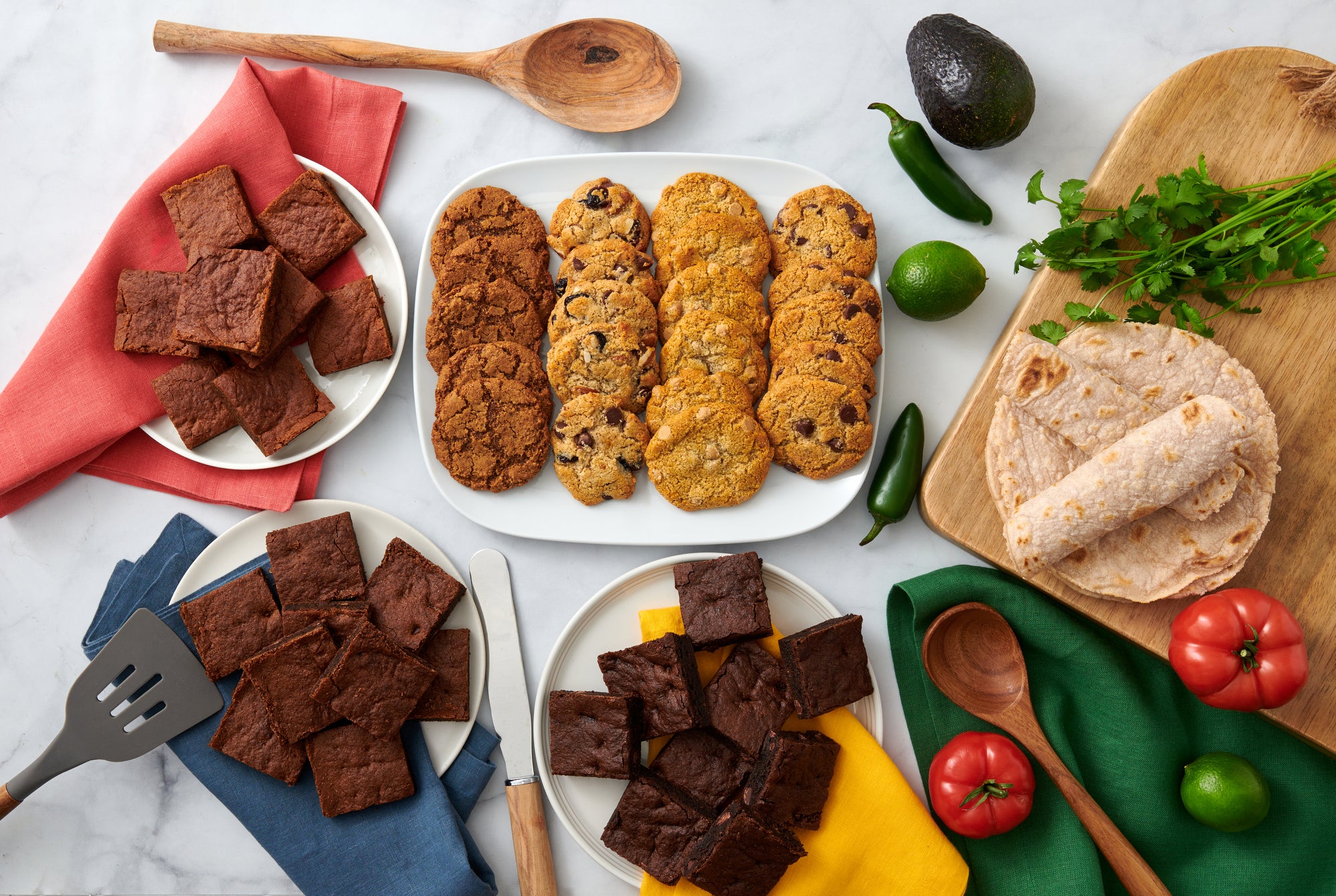 A decorated table with 3 piles of different flavors of brownies on 3 plates, a rectangle plate with rows of 4 kinds of cookies, and some tortillas on a wooden cutting board with a spoon, cilantro, limes, jalapeño, cilantro and tomatoes around them.