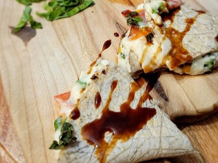 A caprese quesadilla drizzled with pomegranate molasses on a wood cutting board with basil in the background. 
