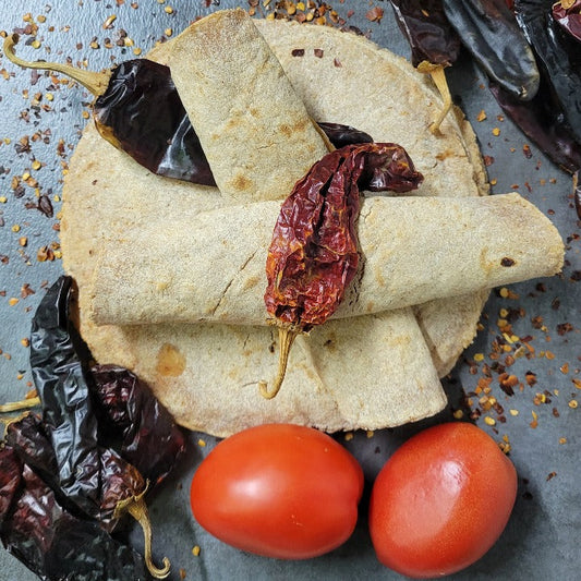 A stack of Southwest Tortillas on a dark gray surface. 2 tortillas rolled up and placed on top in the shape of an X. 2 Dried Red chilies lay on top. Red chiles lay around the tortillas with 2 tomatoes at the bottom.