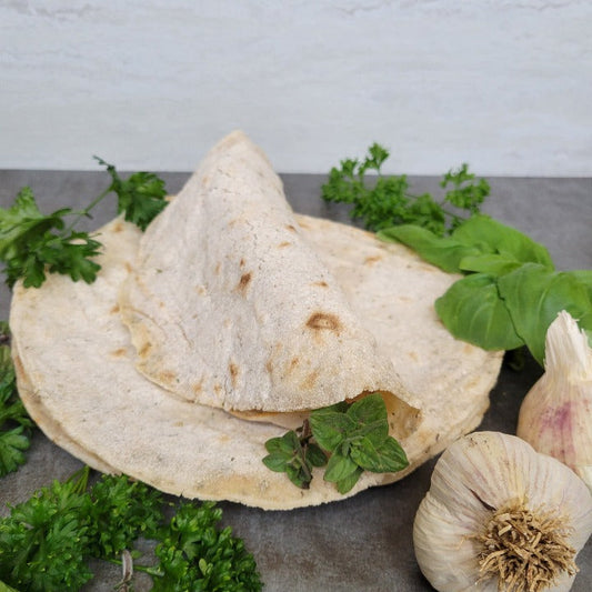 2 garlic and herb tortillas laying flat on a dark gray surface. One folded tortilla on the top with basil and oregano leaves at the opening. Parsley, oregano and garlic bulbs are spread around the tortillas.