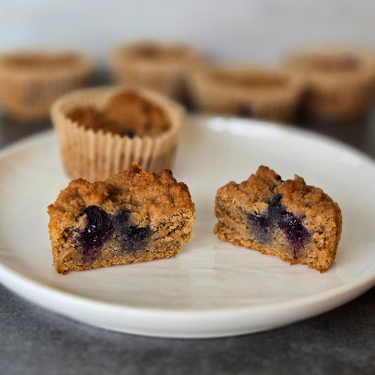 A white plate with 2 blueberry cupcakes, and one cut in half with 4 blueberry muffins in the background