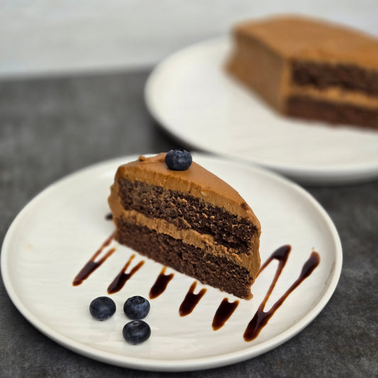 A slice of carob cake on top of a date syrup drizzle with 3 blueberries on a white plate with the whole cake on a white plate in the background.