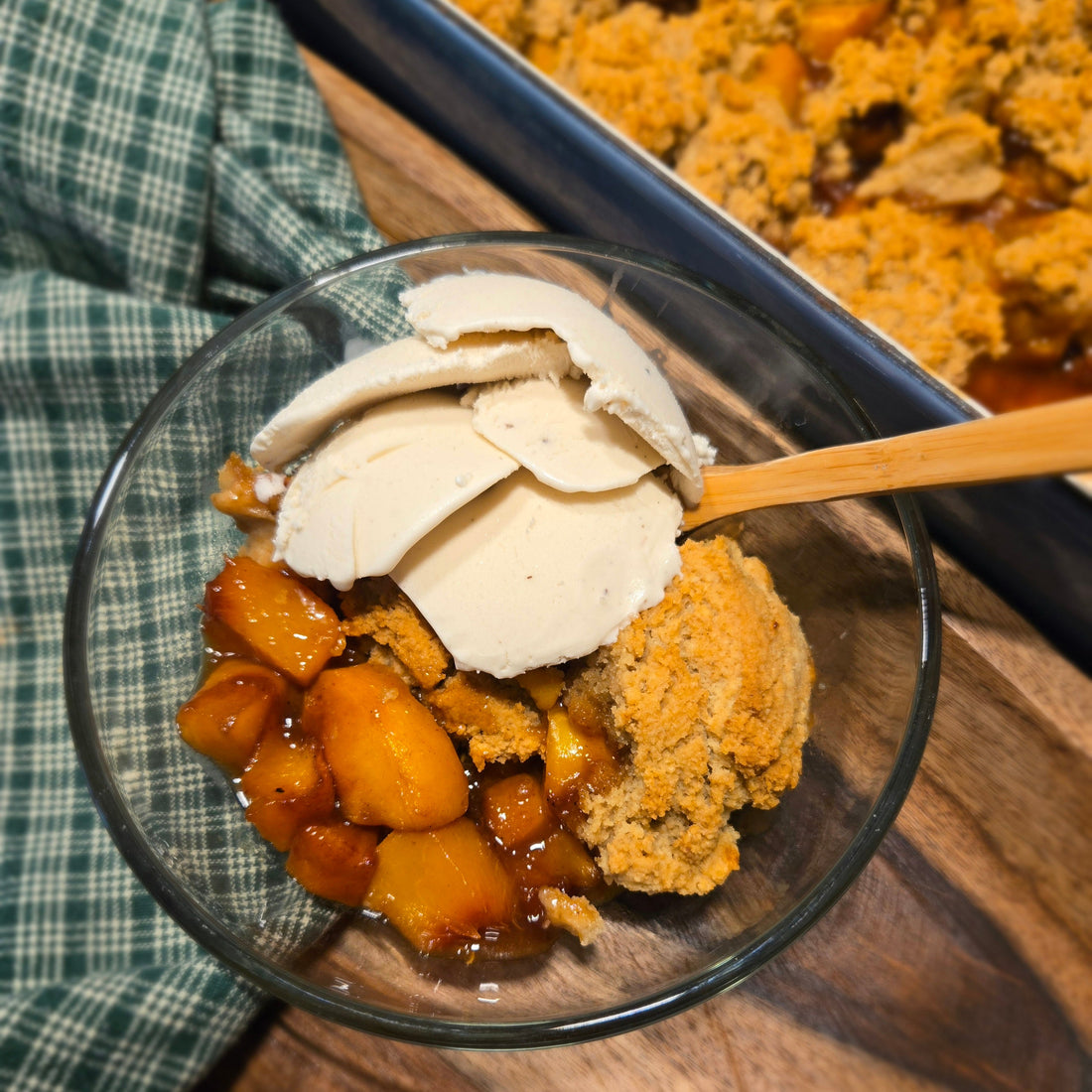 Fresh gluten-free peach cobbler in a glass bowl with vanilla ice cream and a wood spoon sitting on a green checked towel on a wood board with the baked cobbler blurred in the background. 