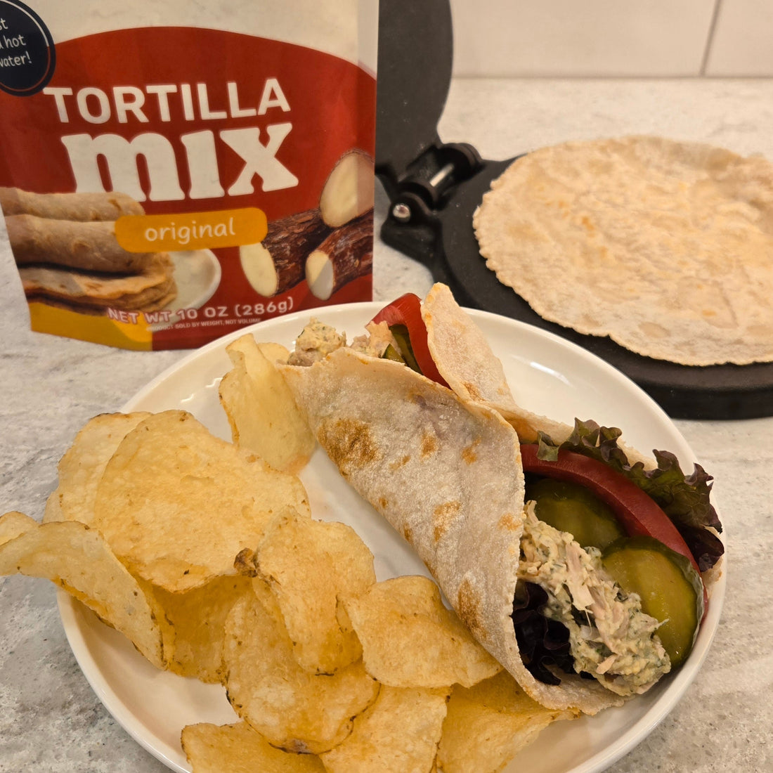 Jackfruit salad wrap on a white plate with potato chips on the left side. A Cassavaberry tortilla mix package in the background on the left, and a tortilla press with a tortilla on it on the right.