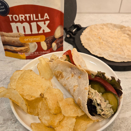 Jackfruit salad wrap on a white plate with potato chips on the left side. A Cassavaberry tortilla mix package in the background on the left, and a tortilla press with a tortilla on it on the right.