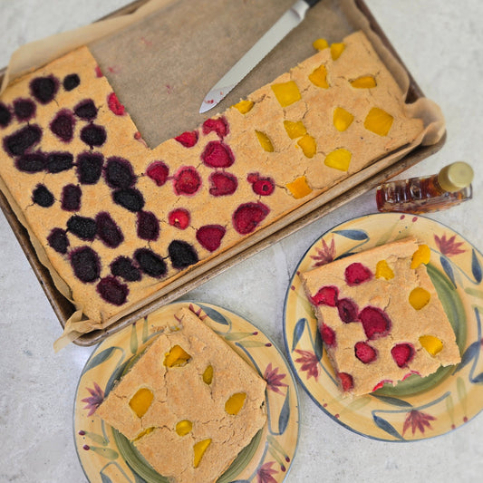 Sheet pancake with blackberries, raspberries and mango. Two square slices on two plates with a bottle of maple syrup to the right.