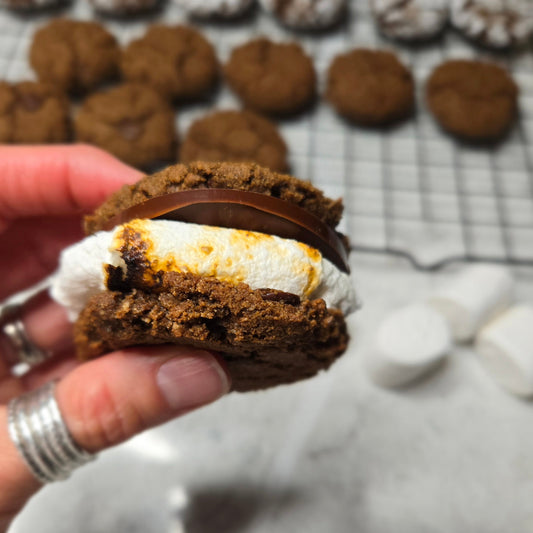 Gluten Free smore made with brownie cookies made from Cassavaberry's Chocolate Chip Brownie Mix. A cooking rack of brownie cookies sits in the background.  