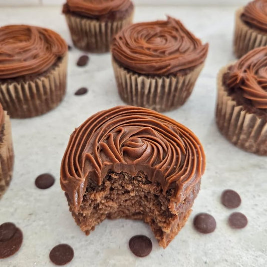 7 frosted carob cupcakes on a white background with bite out of one cupcake with sprinkled carob chips around. 