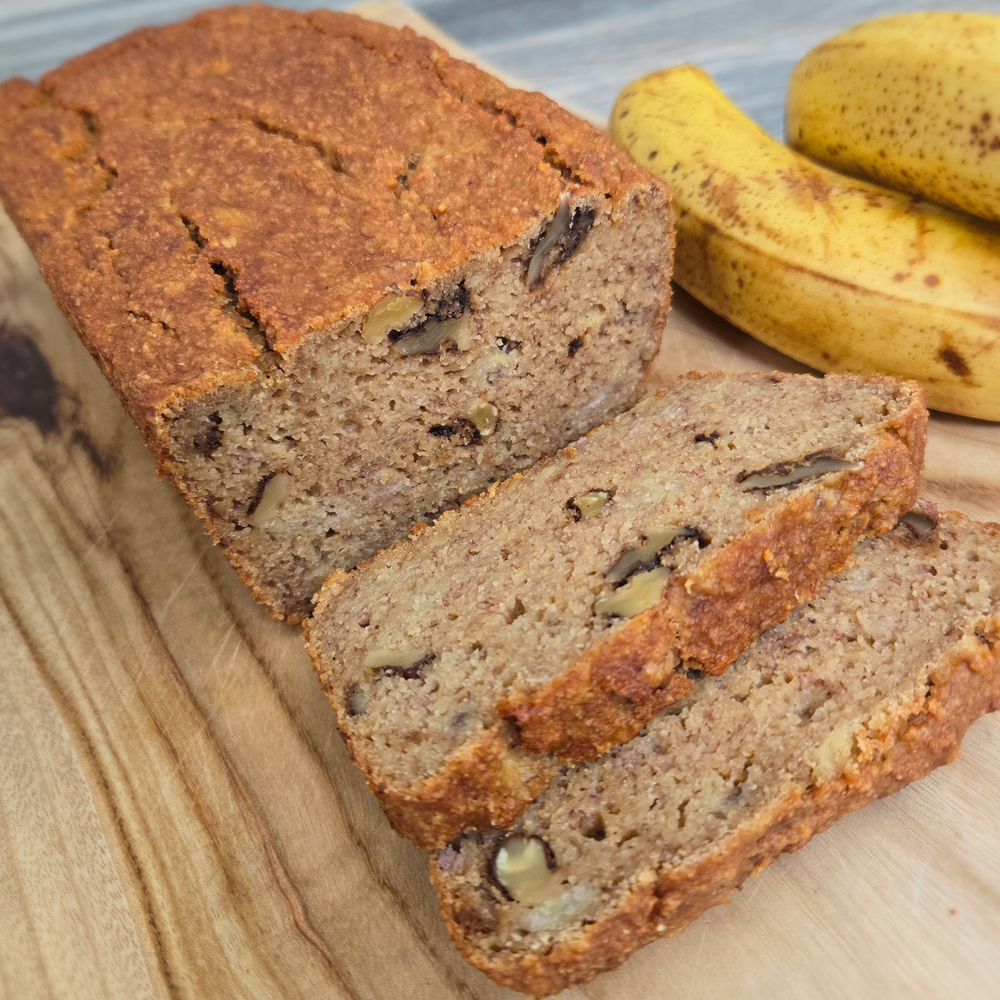 Loaf of banana bread with slices cut, on a wooden surface with bananas in the background.