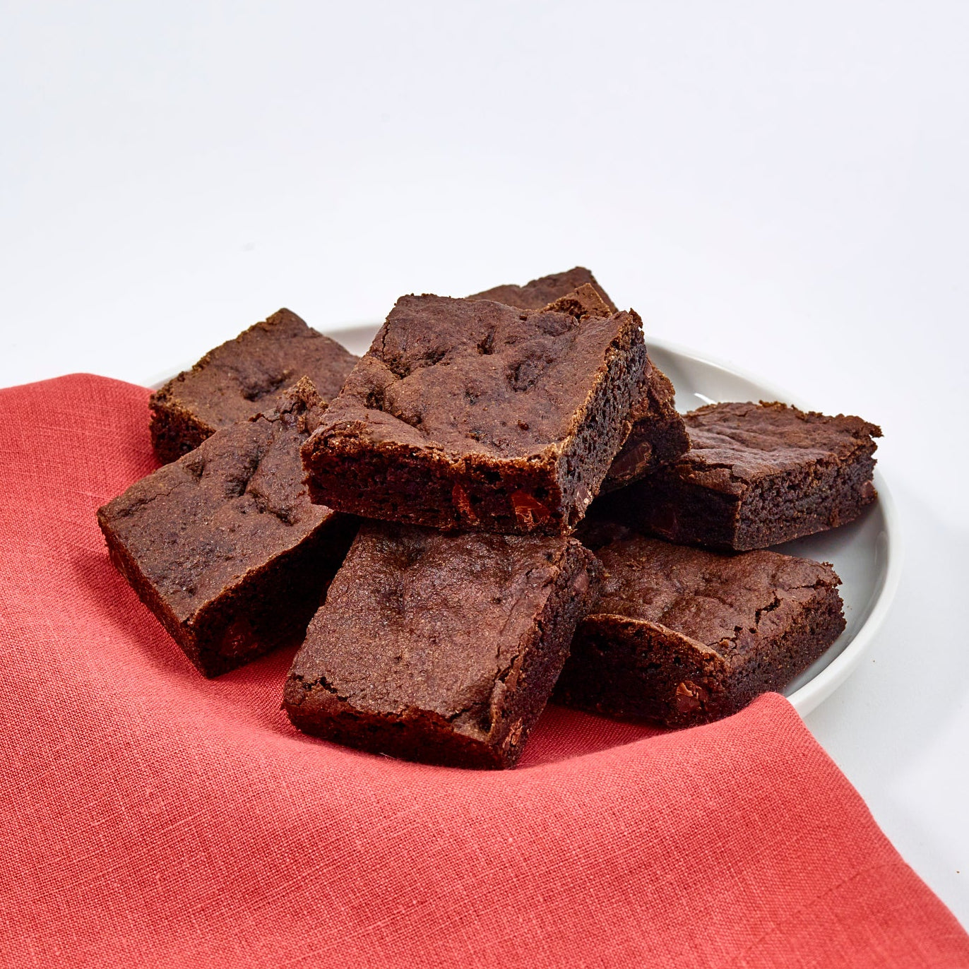 Baked Cassavaberry Chocolate Chip Brownies stacked on a white plate half covered in a red napkin with a white background.