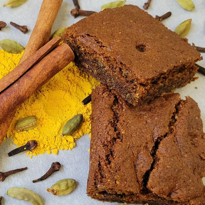 Pumpkin spice brownies stacked with pumpkin powder, cinnamon sticks, cardamon pods, cloves around the brownies on a white background.