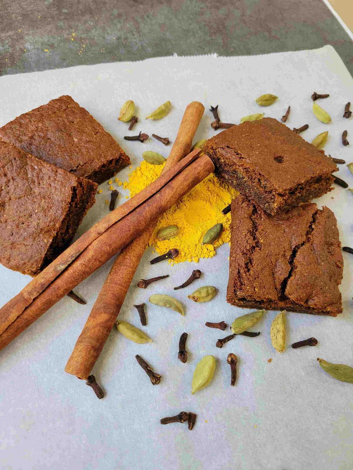 Pumpkin Spice Brownies arranged on parchment paper with cinnamon sticks, whole spices, and a  pile of pumpkin powder.